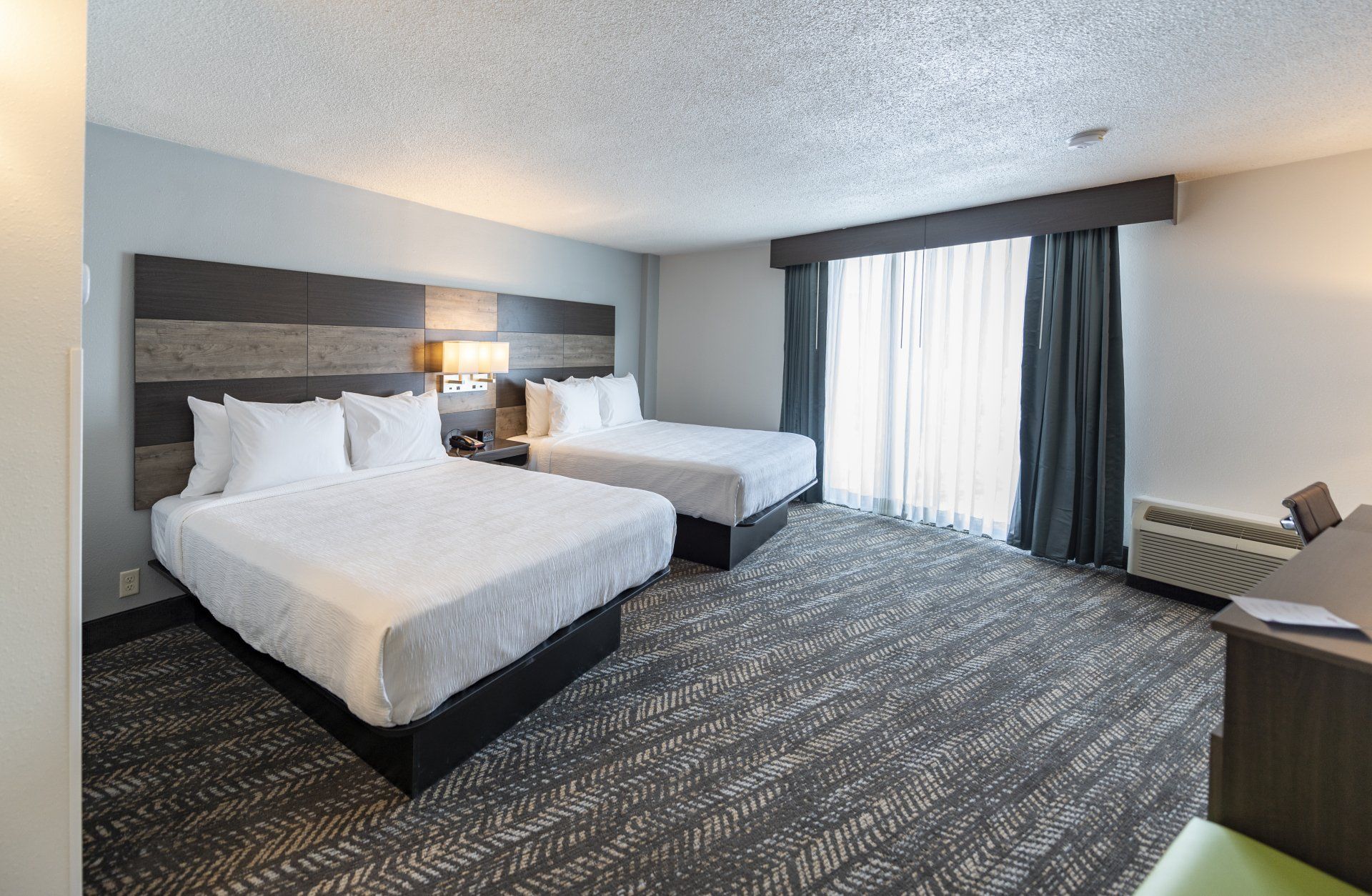 Hotel room with two queen beds, gray carpet, and a large window with curtains.