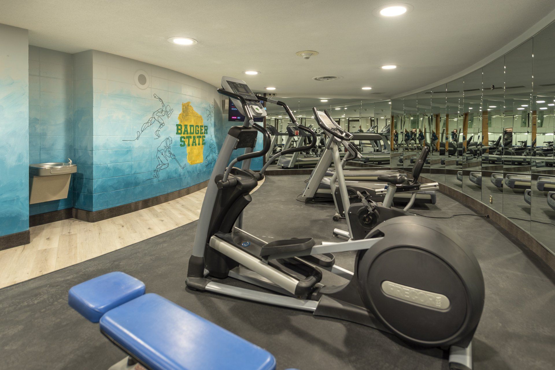 Gym interior with elliptical machine, weights, and treadmills. Blue and white mural.