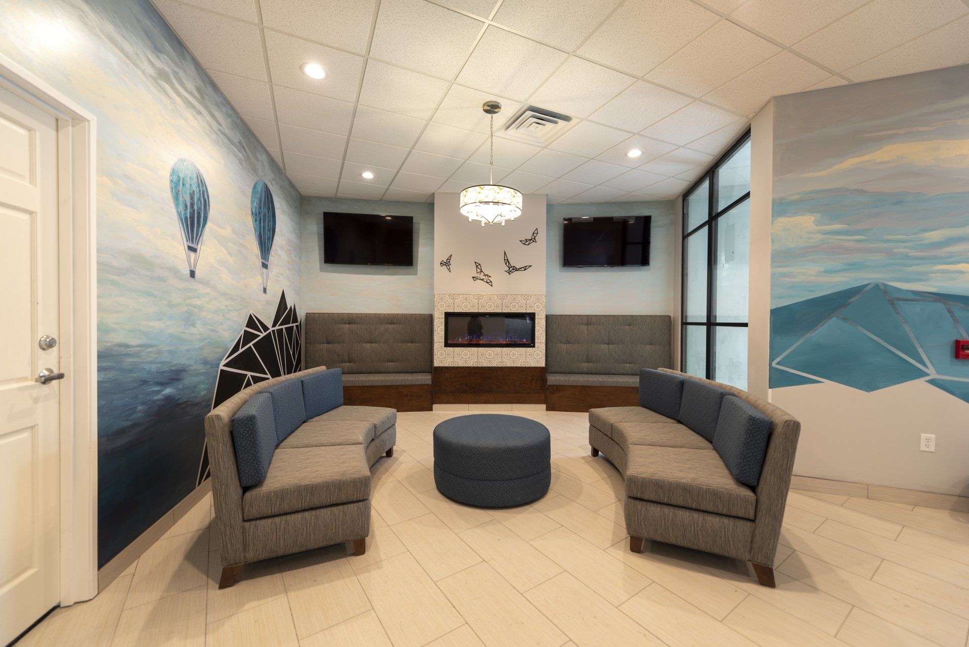 A waiting room with two blue sofas, a round ottoman, and a fireplace, against a mural of mountains and hot air balloons.