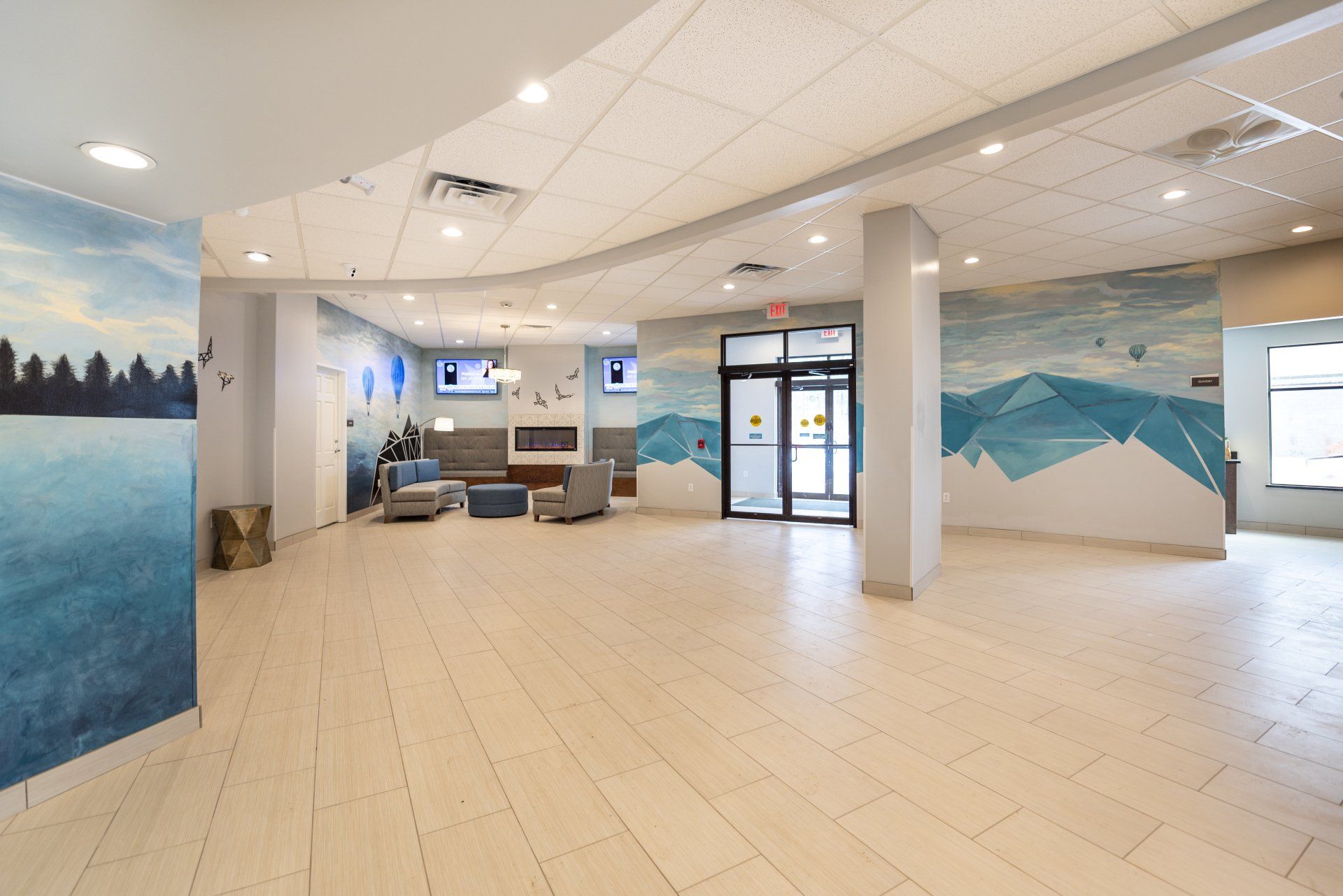 Bright lobby with light wood floors, landscape murals, and seating near a fireplace and entrance doors.