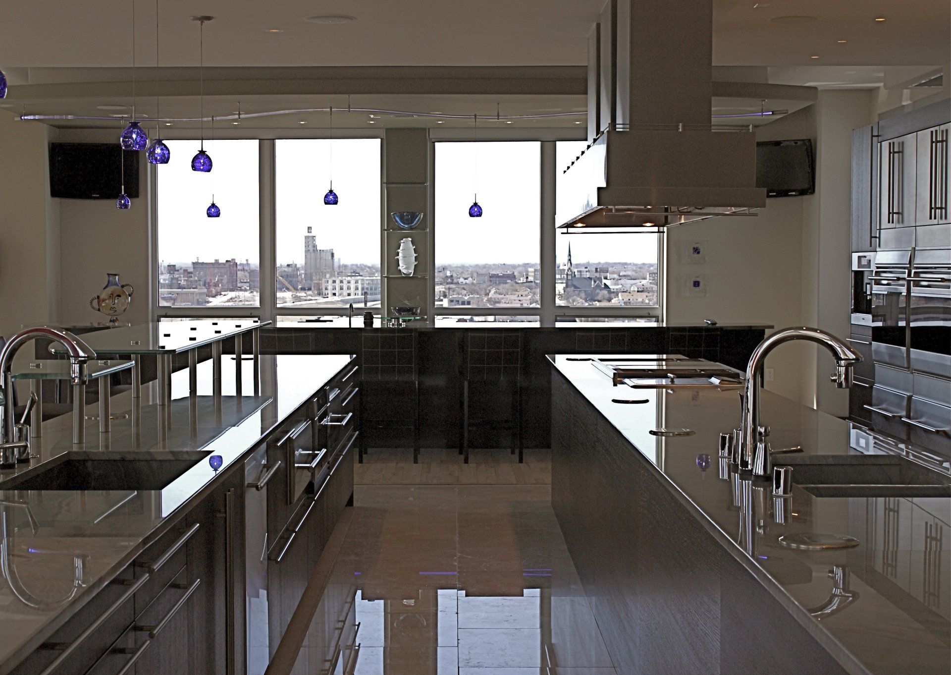 Modern kitchen with two islands, large windows overlooking a city skyline, and blue pendant lights.