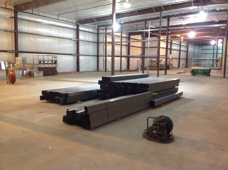 Interior view of a large warehouse under construction, metal beams and equipment on concrete floor.