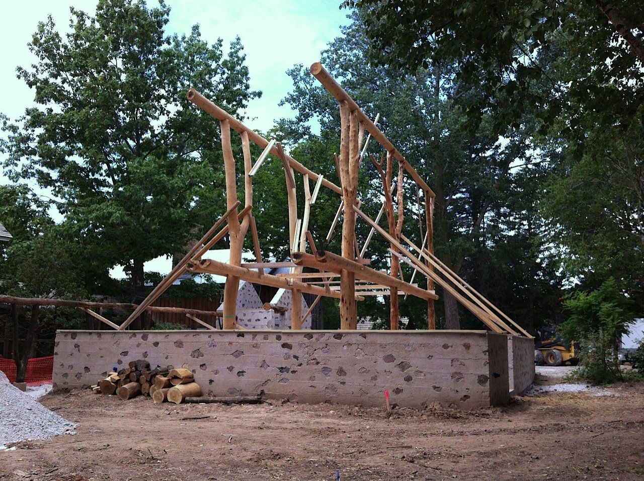 Wooden framework of a building under construction on a concrete foundation, outdoors with trees in the background.