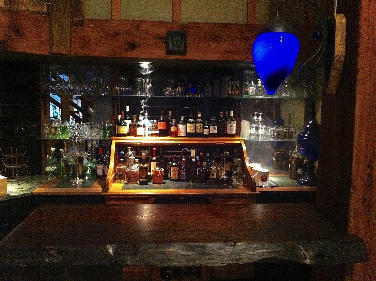 Wooden bar with liquor bottles and glassware; blue glass light hangs overhead.