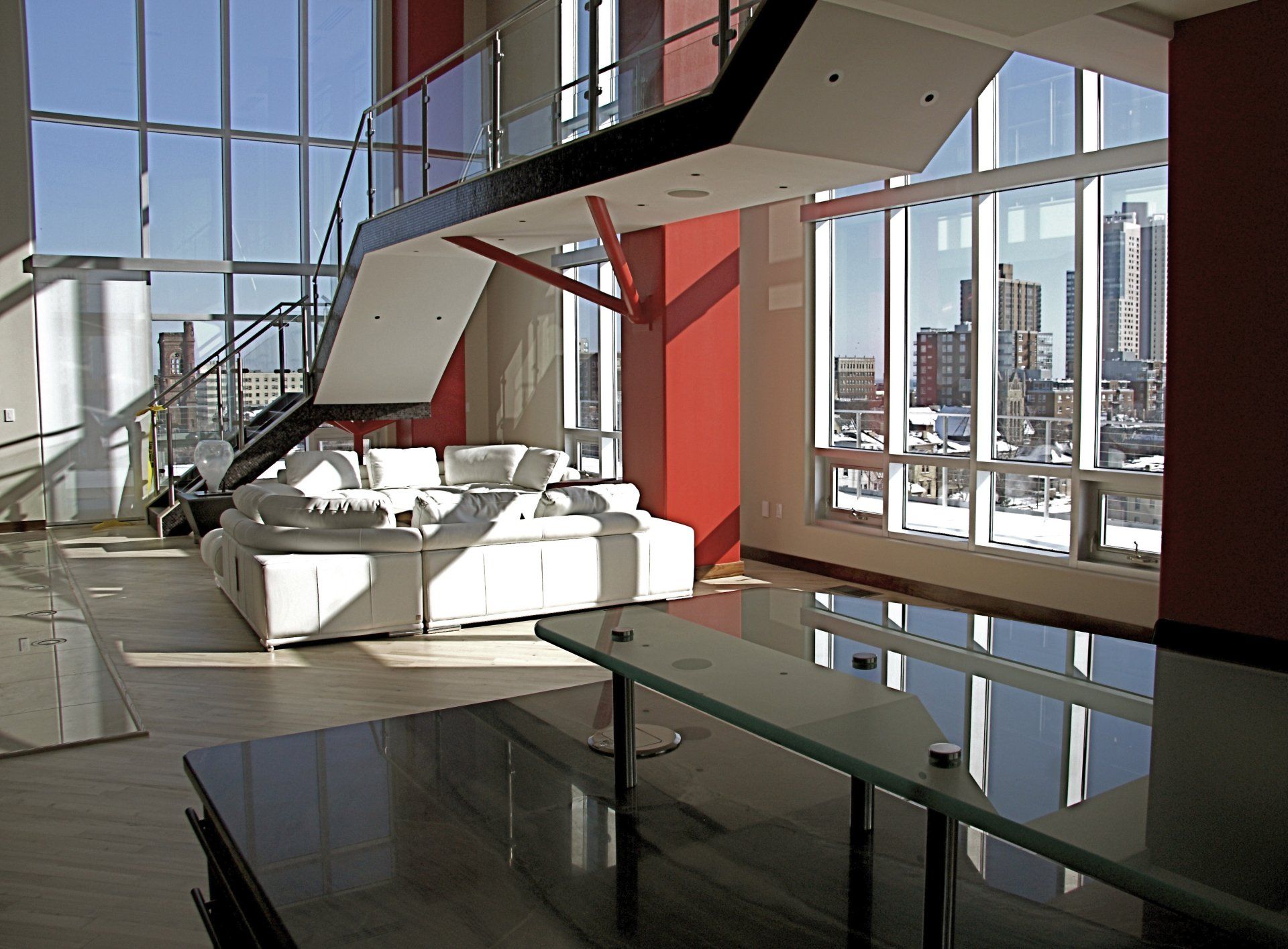 Modern, open-plan apartment interior with floor-to-ceiling windows overlooking a city skyline. Features a floating staircase and white sofa.