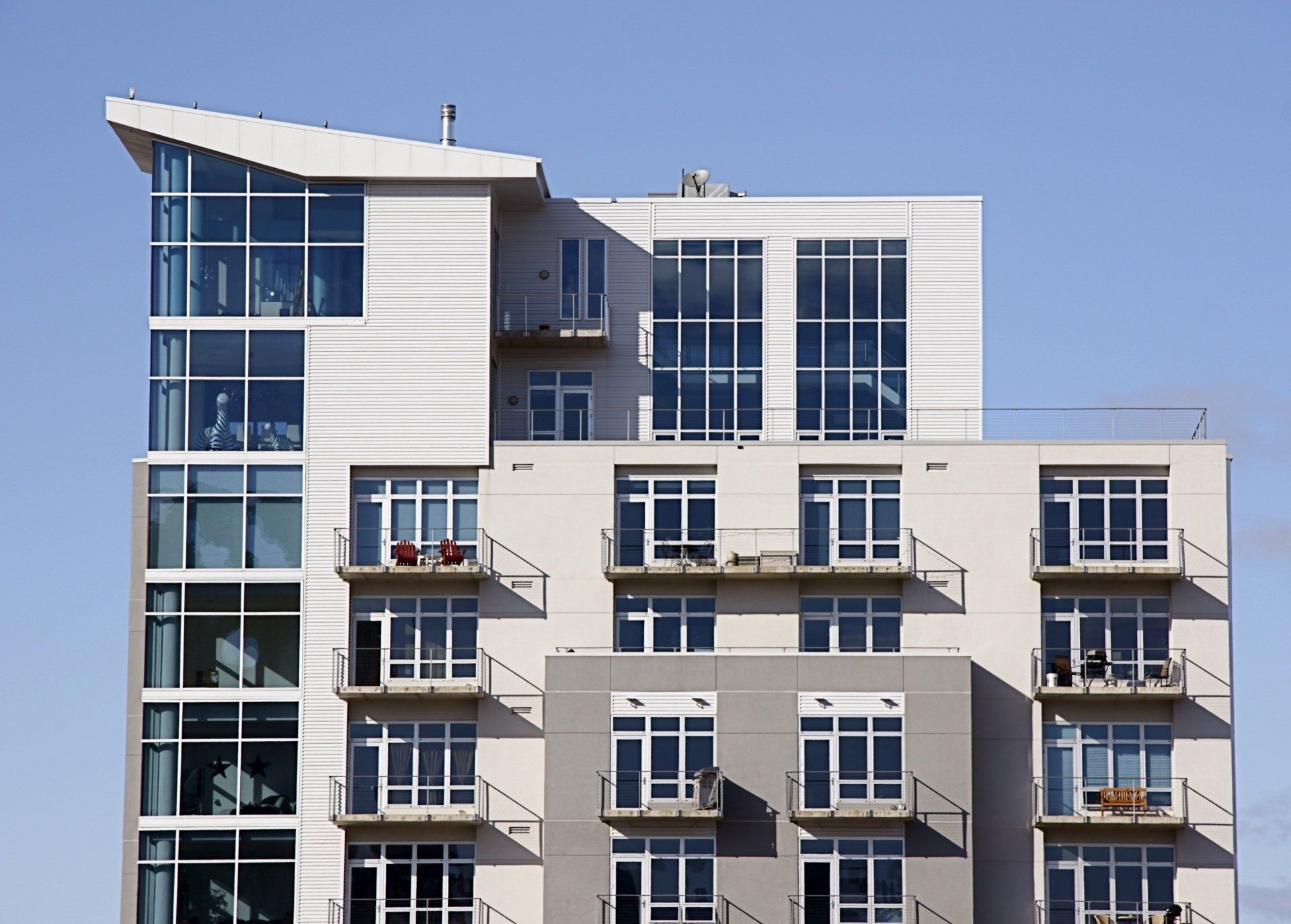 Modern multi-story apartment building with balconies, glass windows, and white siding against a blue sky.