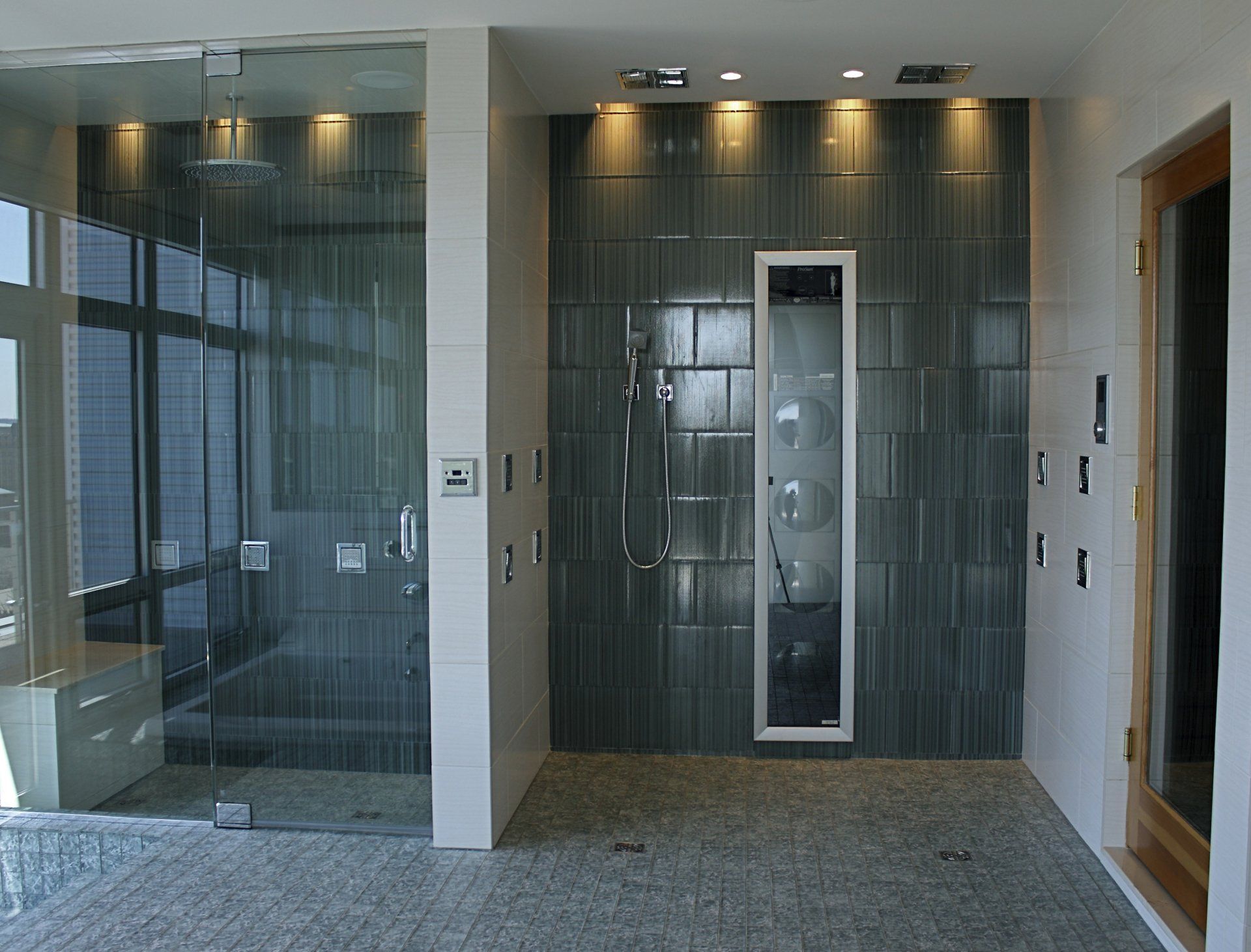 Modern bathroom with two showers, one glass-enclosed. Gray tile walls and floor. Recessed lighting.