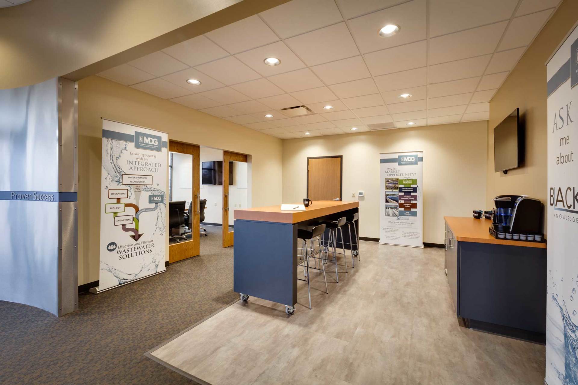 An office break room with a long table, chairs, coffee station, and two posters.