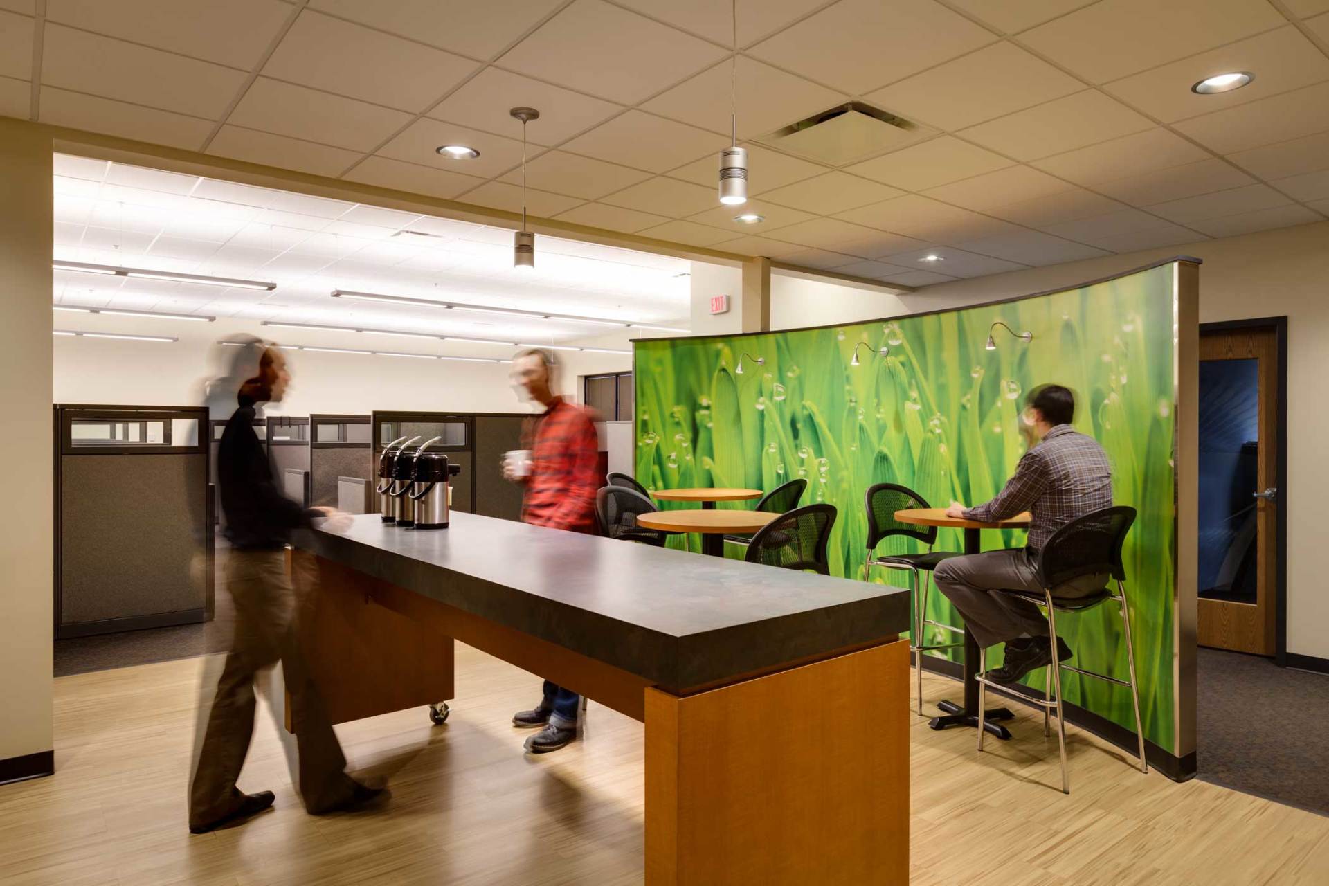 Office break room with people near coffee bar and table with decorative green screen.