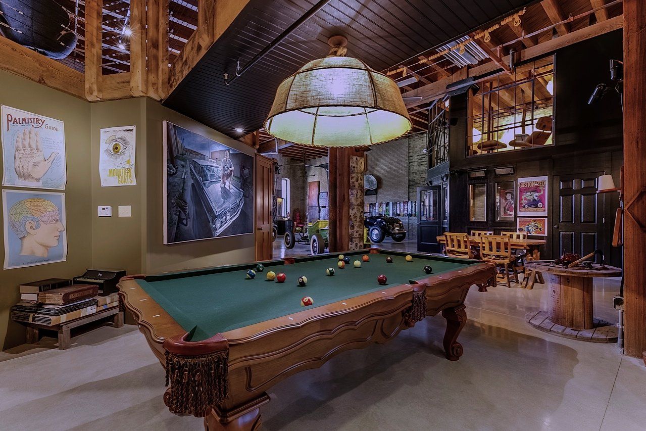 Pool table in a rustic room with wood beams, a large hanging lamp, art on the walls, and a small sitting area.
