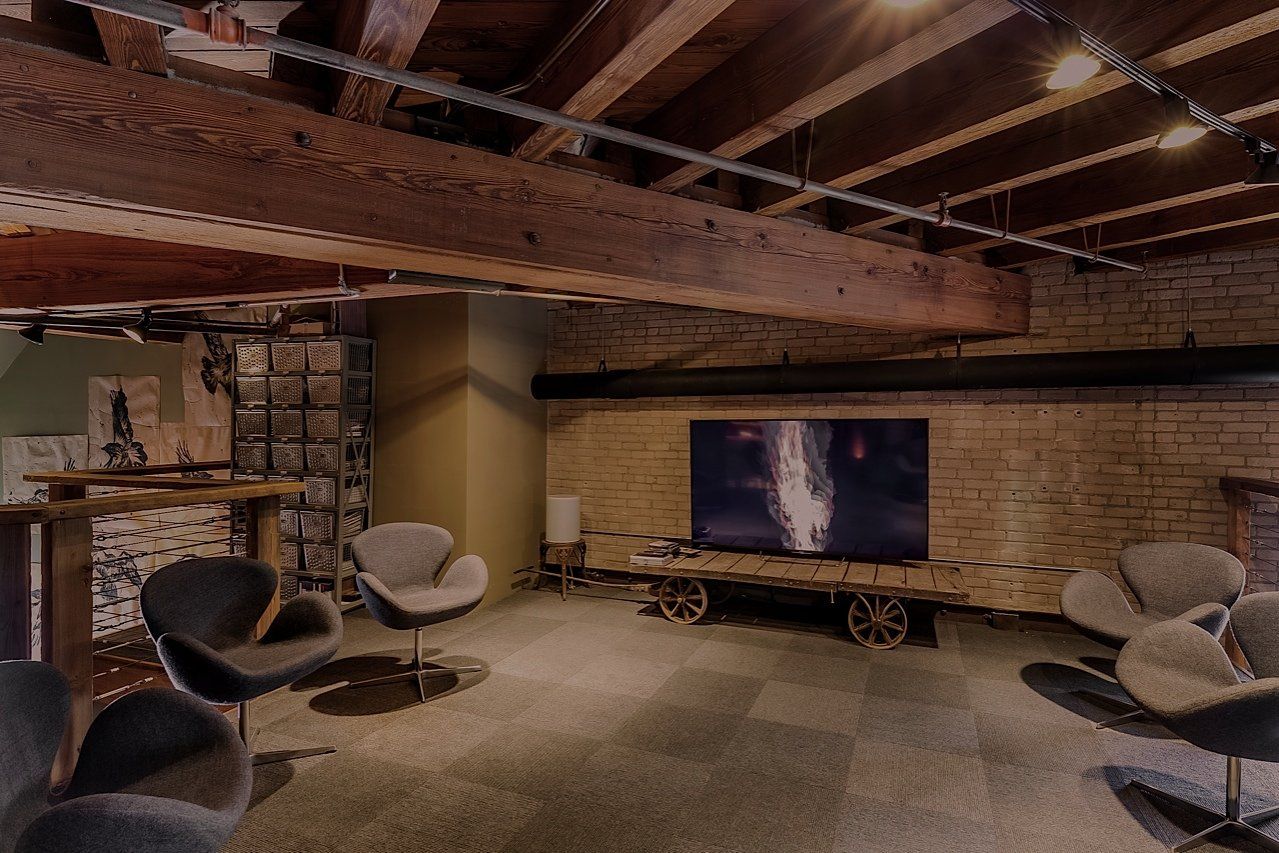 Lounge area with gray chairs, a TV, and exposed wood beams. A brick wall and a table are also visible.