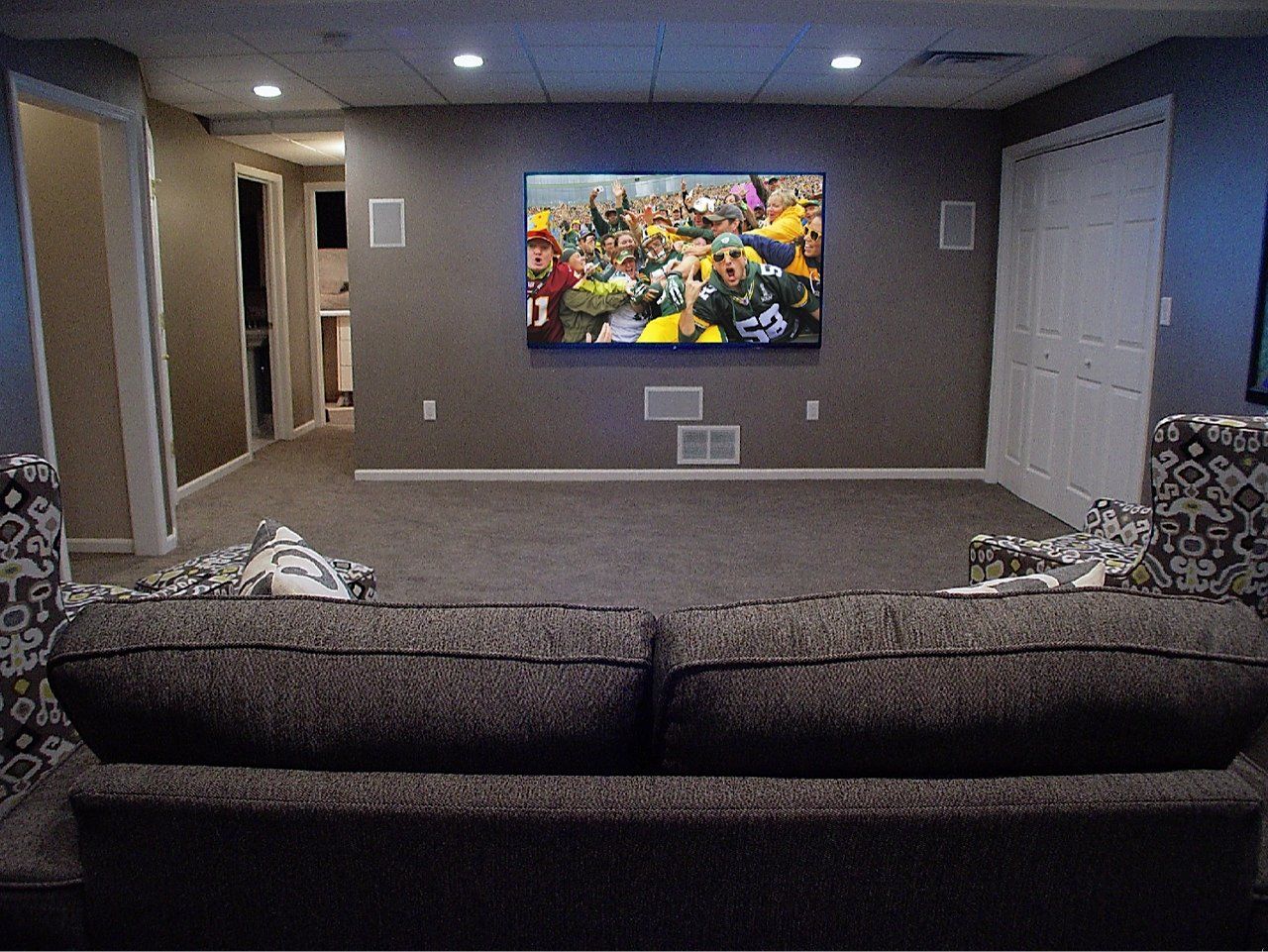 A living room with a large TV showing a sporting event above a gray couch, flanked by patterned chairs.