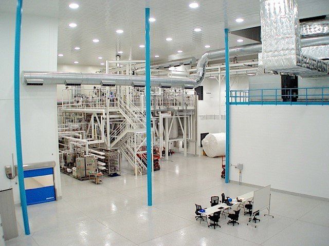 Large, white industrial space with complex machinery, piping, and a small conference table in the foreground.