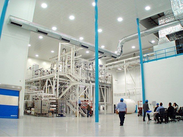 Large industrial facility with complex machinery; several people in hard hats and suits. White and blue color scheme.