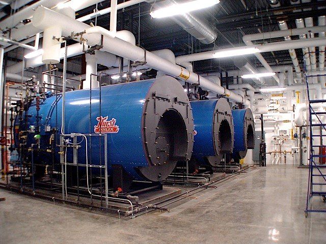 Blue industrial boilers in a room with pipes and metal scaffolding.