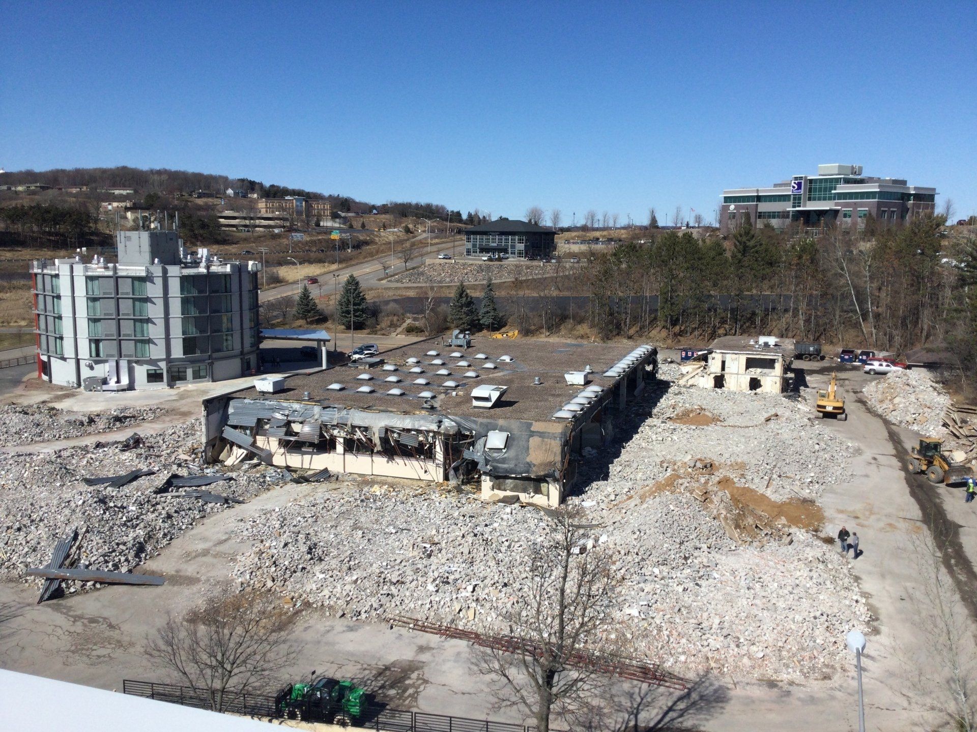 Demolition site with rubble and partially destroyed building; other buildings and hills in background.