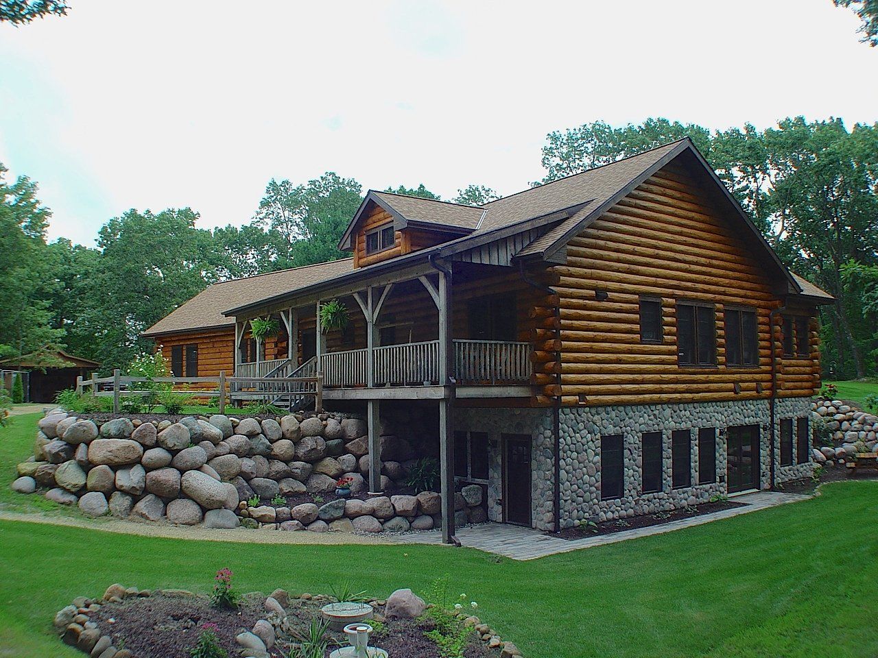 Log cabin house with stone foundation and wrap-around porch, set on a green lawn.