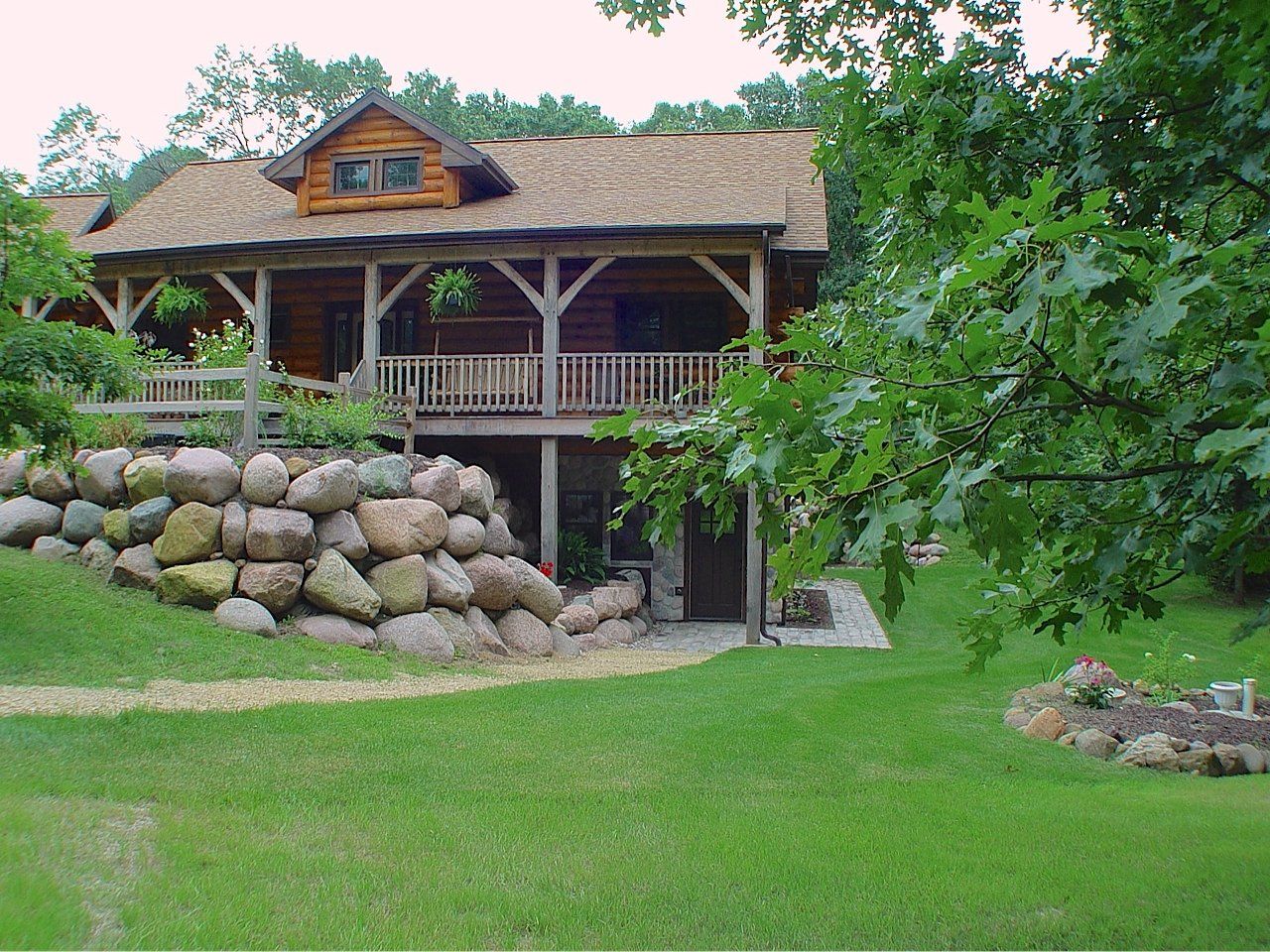 Log cabin with a thatched roof, surrounded by green grass, trees, and a stone wall.