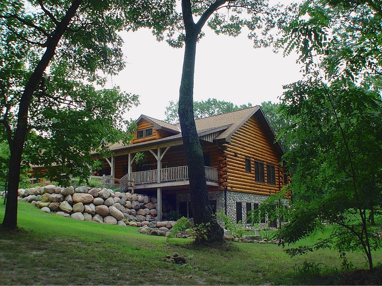 Log cabin with wooden siding, stone foundation, and a wraparound porch, nestled in a wooded setting.