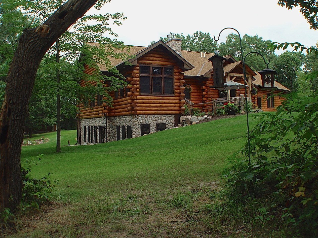 Log cabin house on a grassy hill, with stone foundation, wooden roof, surrounded by trees.