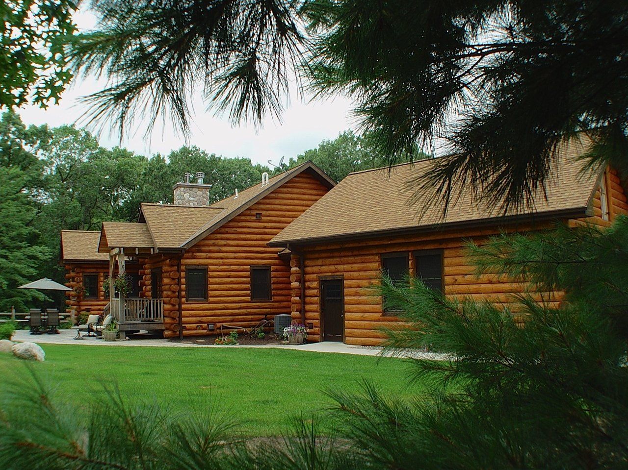 Log cabin with a shingled roof and stone chimney, set in a grassy yard, with surrounding trees.