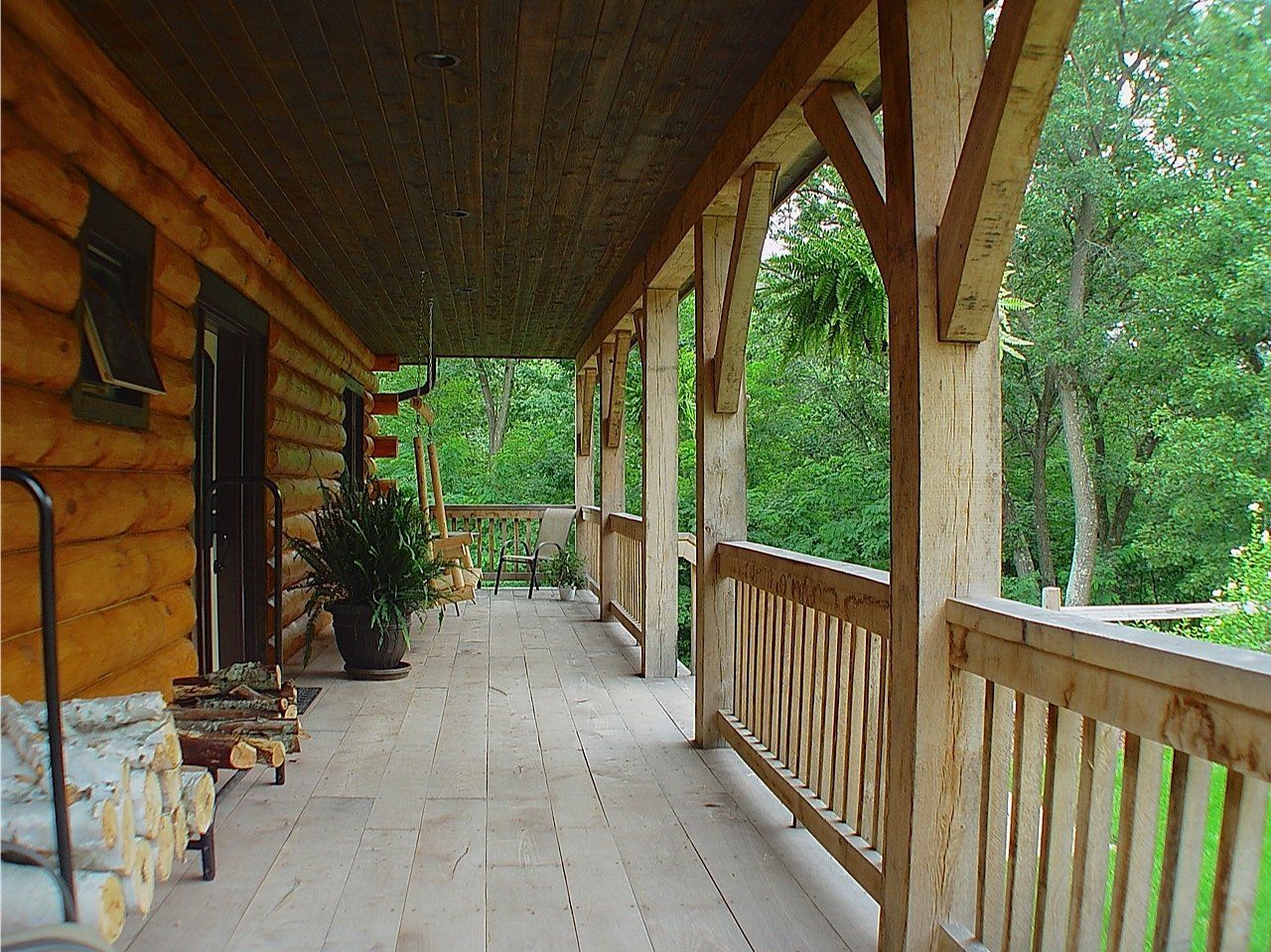 Log cabin porch with wooden railing, surrounded by trees and a stack of firewood.