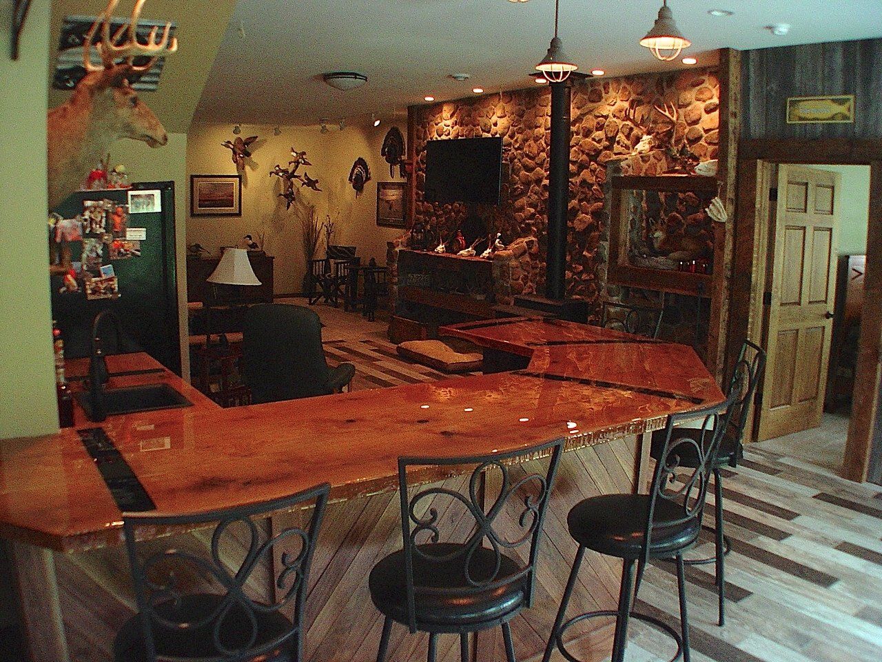 Rustic home bar with wooden counter, stone fireplace, mounted deer head, and bar stools.