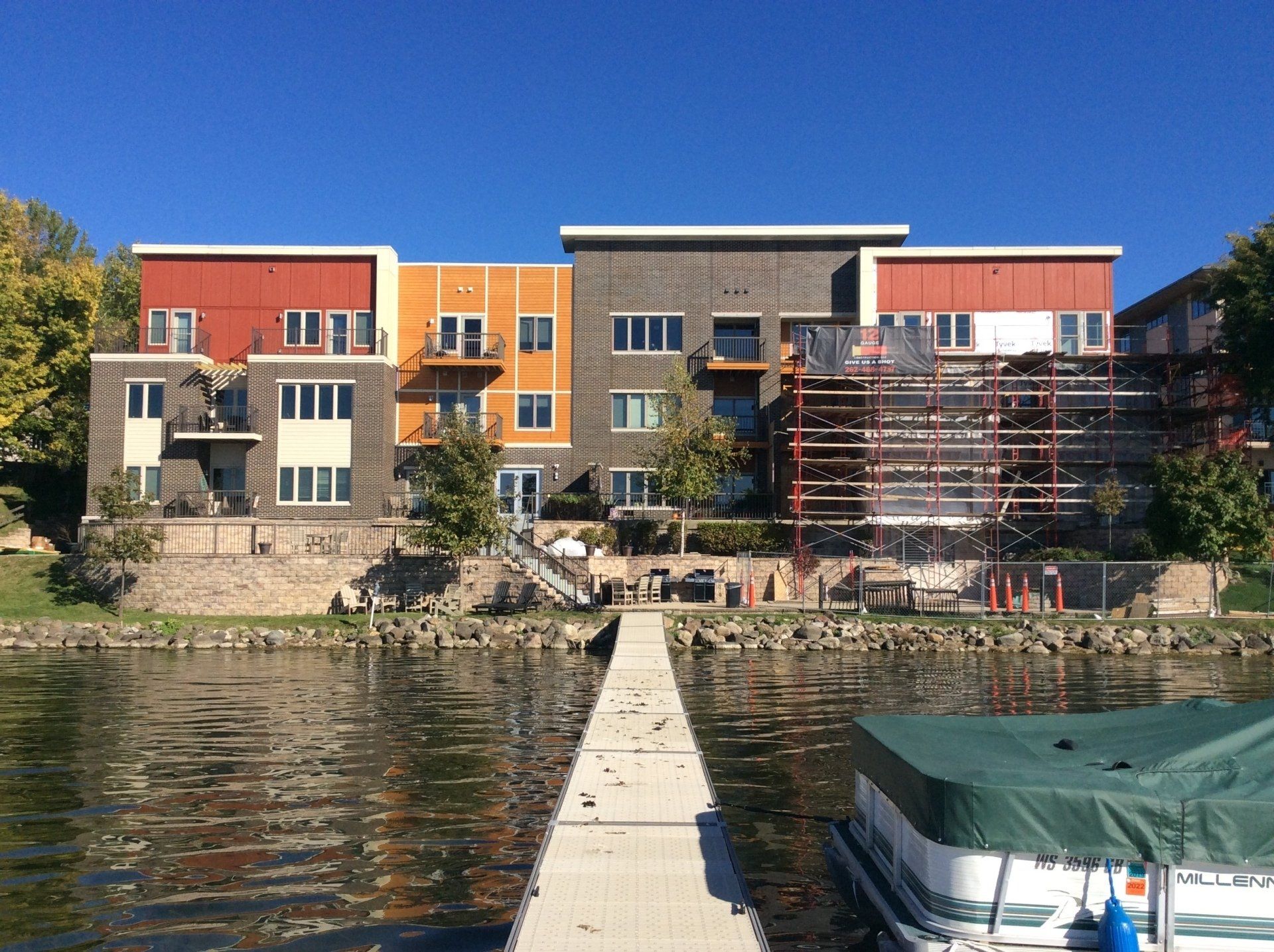 Multi-story waterfront building with dock and boat on a sunny day. Building has brick and red accents.