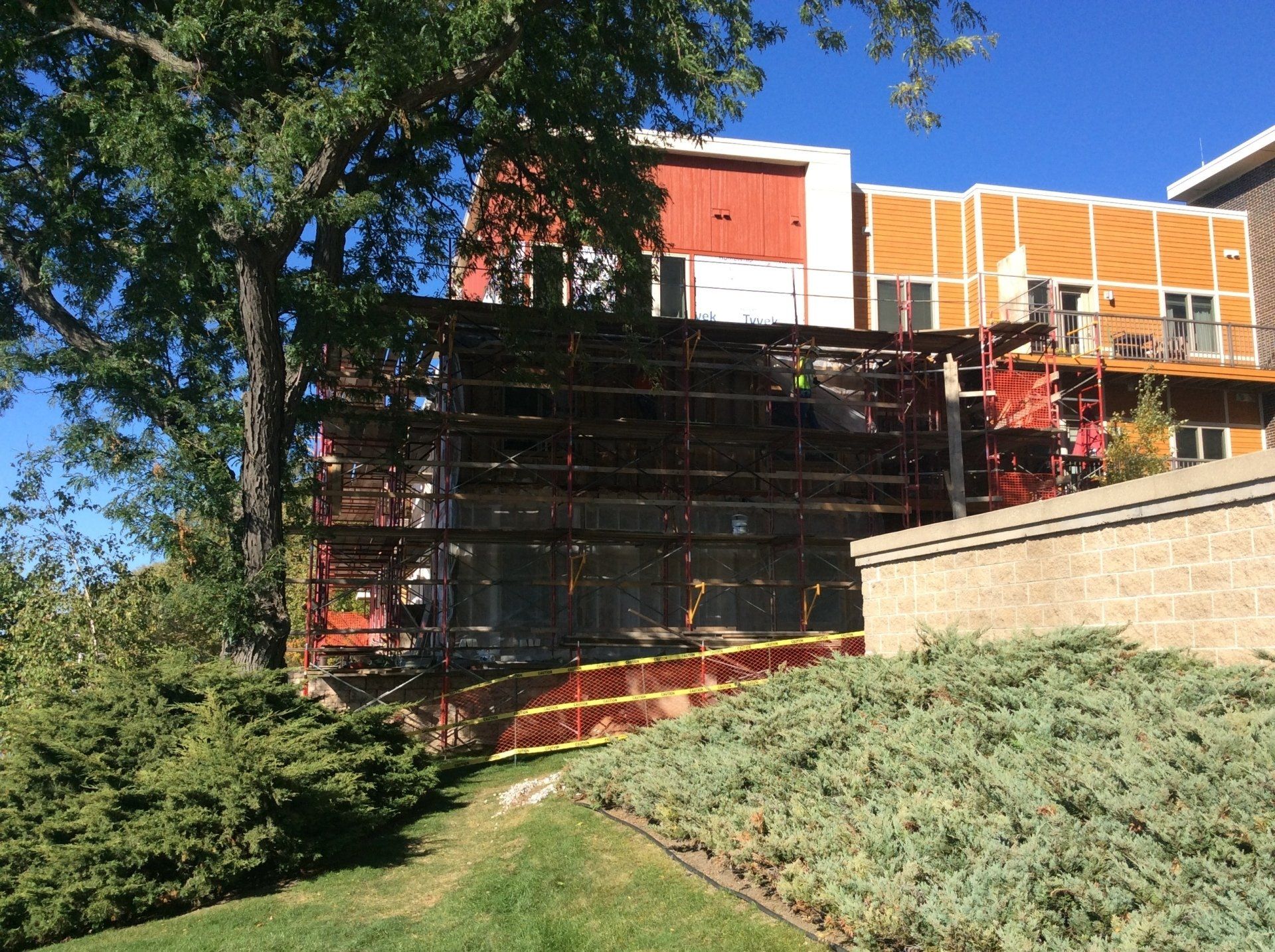 Building construction site, metal scaffolding, and orange and tan building, trees, and green bushes.
