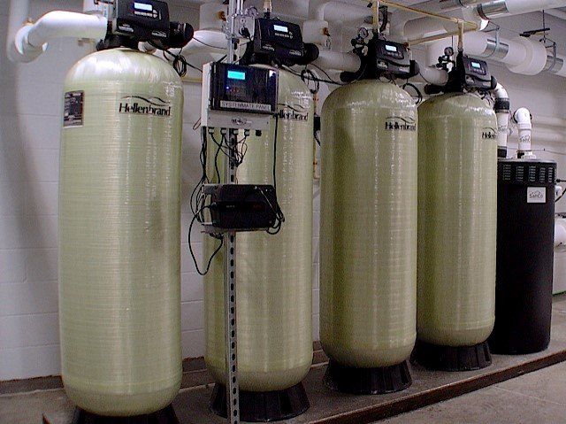 Four tall, cylindrical water filtration tanks with control panels, in a utility room.
