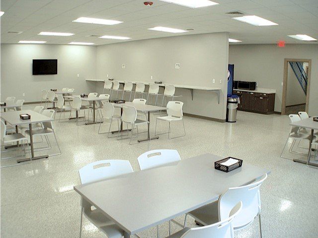 A bright, empty cafeteria with square tables, white chairs, and a TV on the wall.