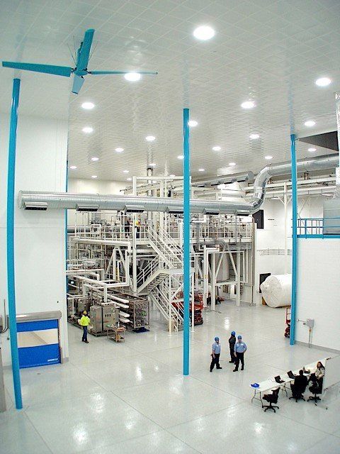 Large factory interior with tall equipment, workers, and a large overhead fan.