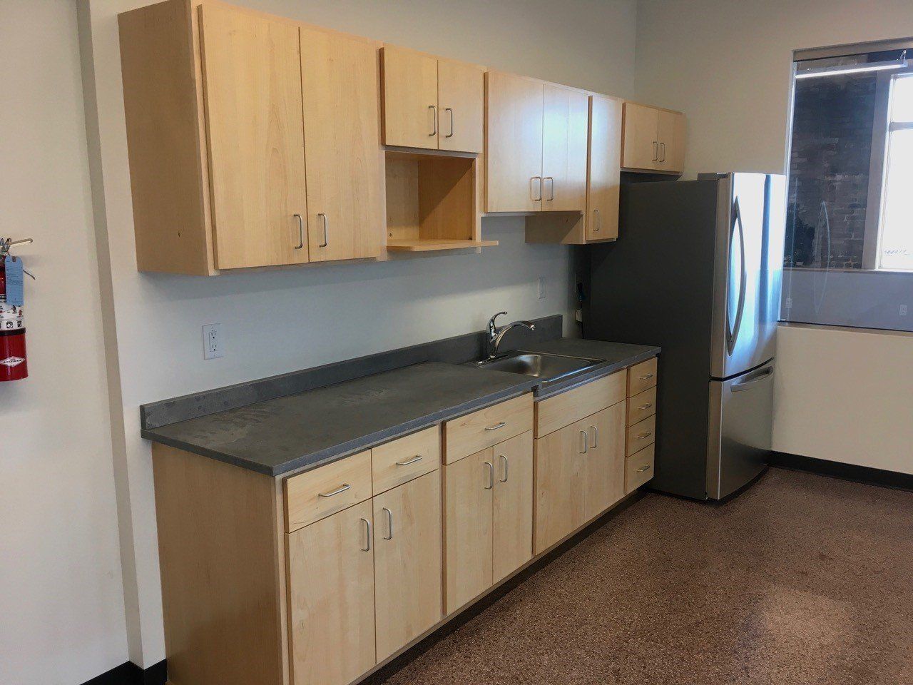 Kitchen with light wood cabinets, gray countertop, stainless steel refrigerator, and a sink.