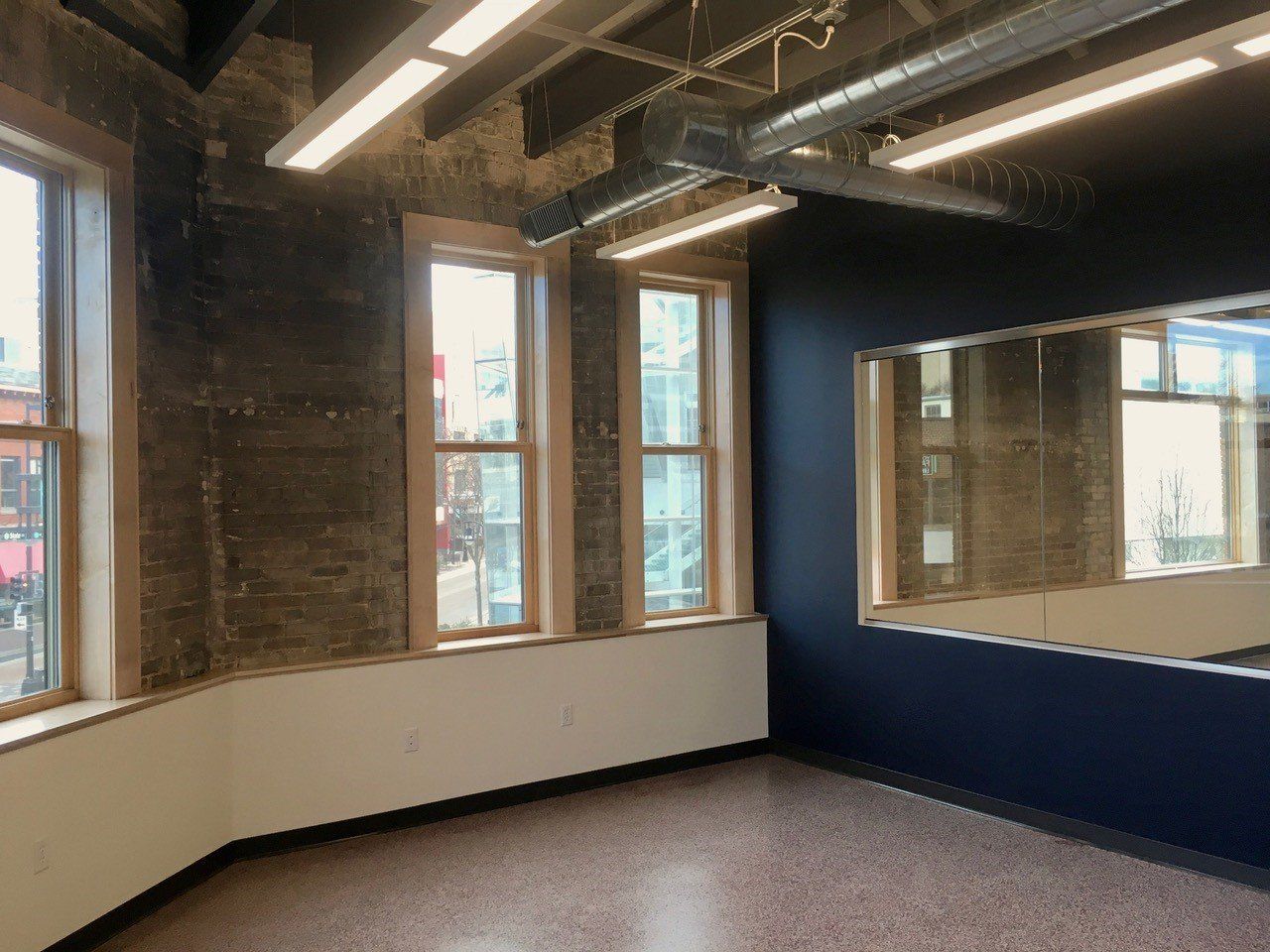 Empty room with large windows, mirror, and dark blue wall. Beige flooring and exposed ceiling beams.