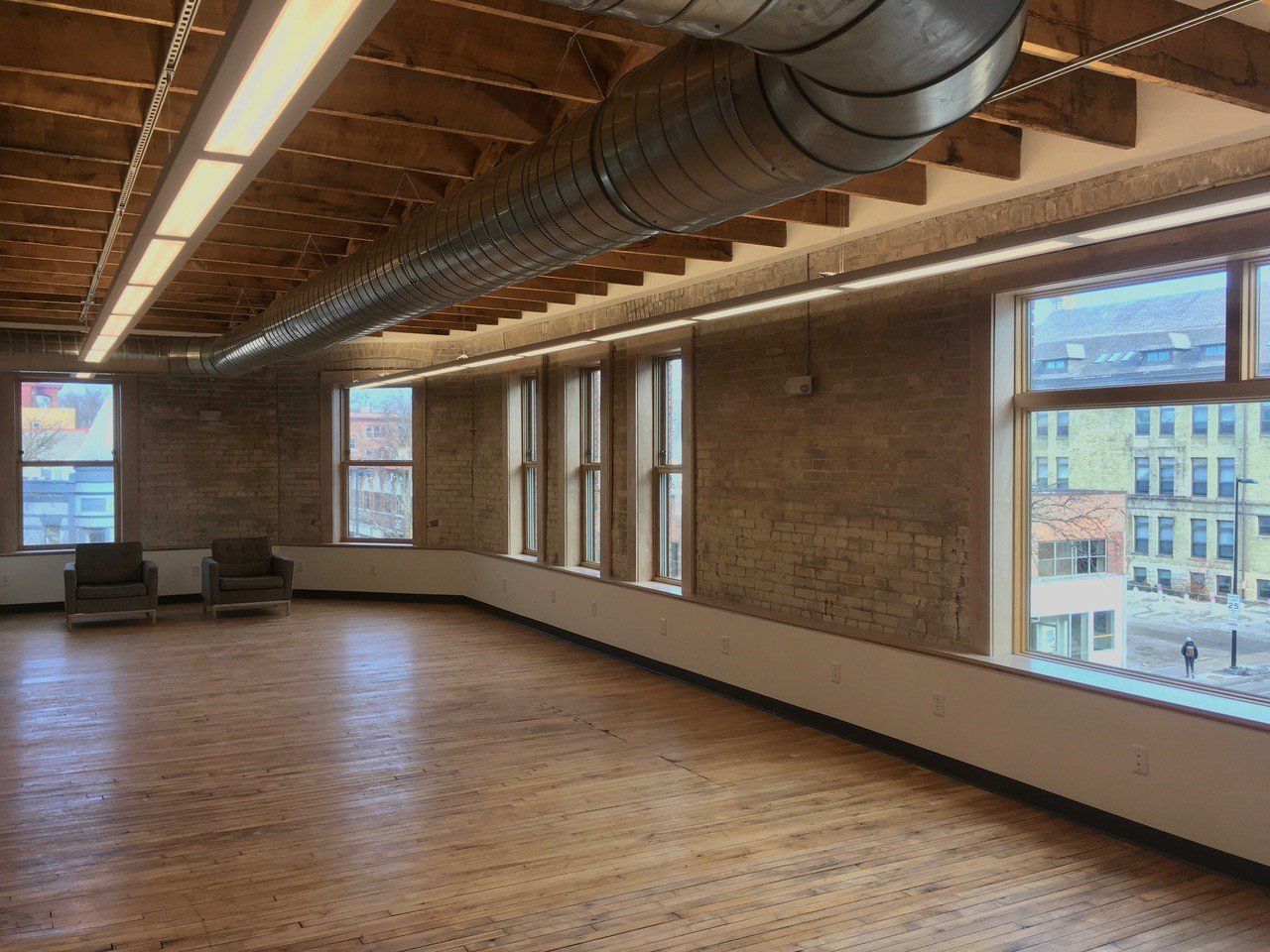 Empty room with wood floor, exposed brick wall, windows overlooking a street, and exposed ductwork.