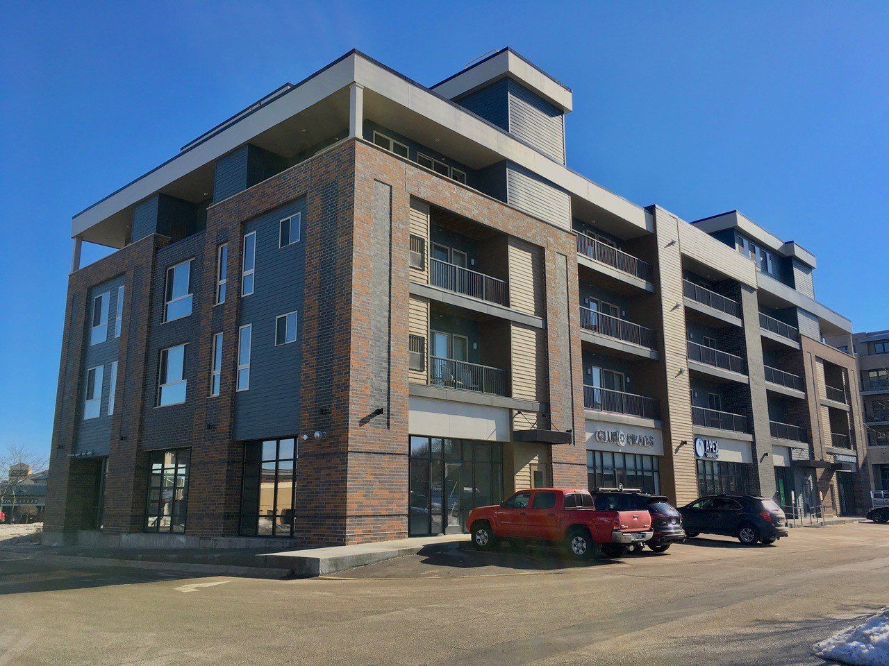 Modern brick and gray building with retail space on the ground floor; cars parked nearby, blue sky.