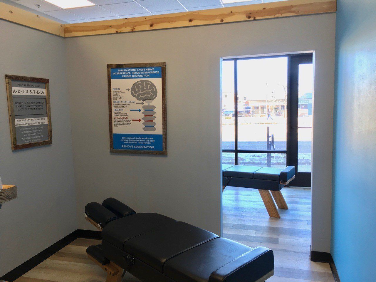 Chiropractor's office: gray walls, treatment table, educational posters, doorway to waiting area with bench.