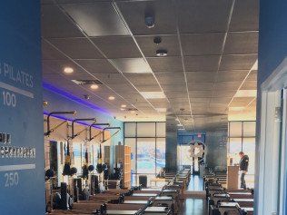 Pilates studio interior with exercise equipment, windows, and person standing. Blue wall, grey ceiling.