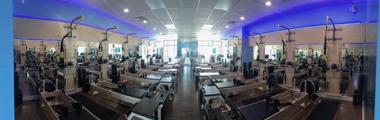 Gym interior with rows of workout machines, blue ceiling lighting, and windows.