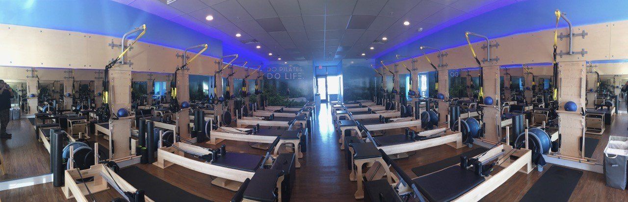 Pilates studio with exercise equipment arranged in rows. Blue and neutral-toned interior with bright lighting.