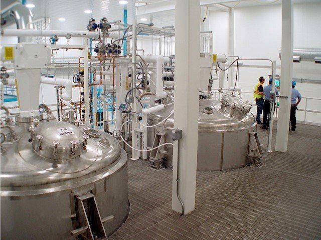 Large industrial tanks and piping in a clean room, two men in the background observing.