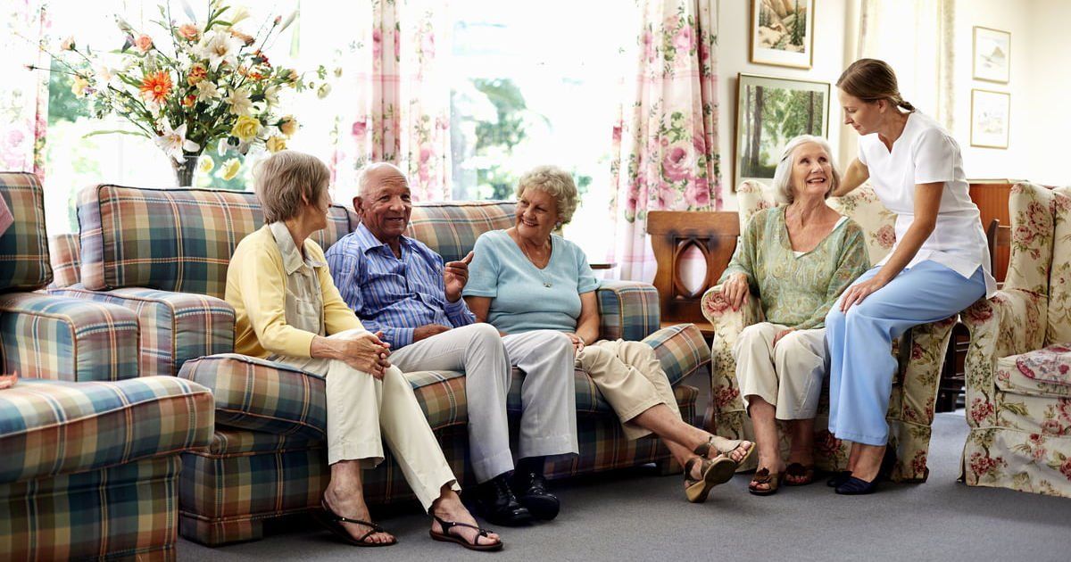 Group of seniors and caregiver sitting on floral sofas in a sunlit room.