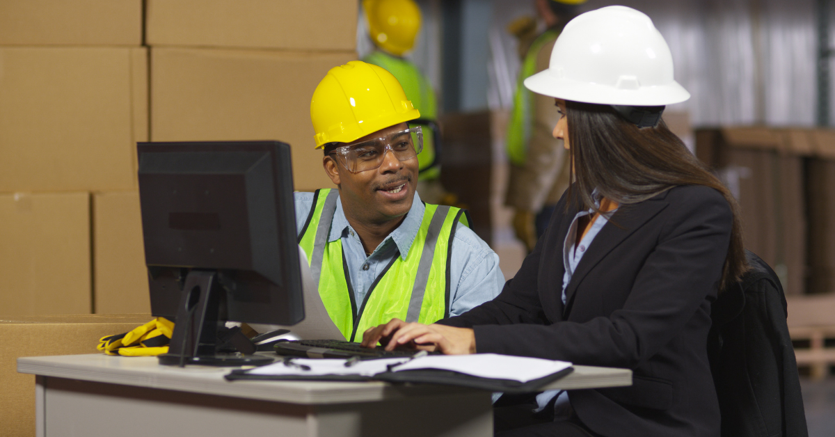 Woman in suit and worker in vest look at computer in warehouse.