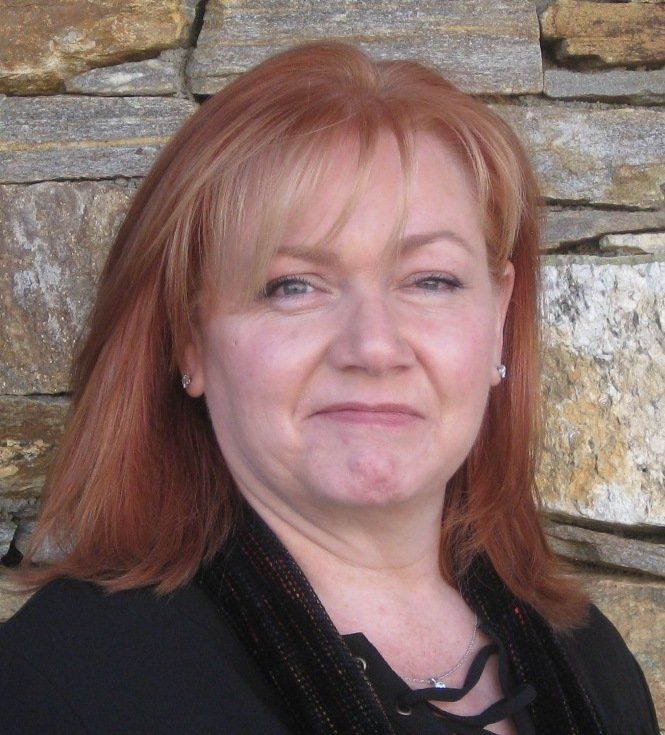 Woman with reddish hair in front of a stone wall, wearing a black top and a slight smile.
