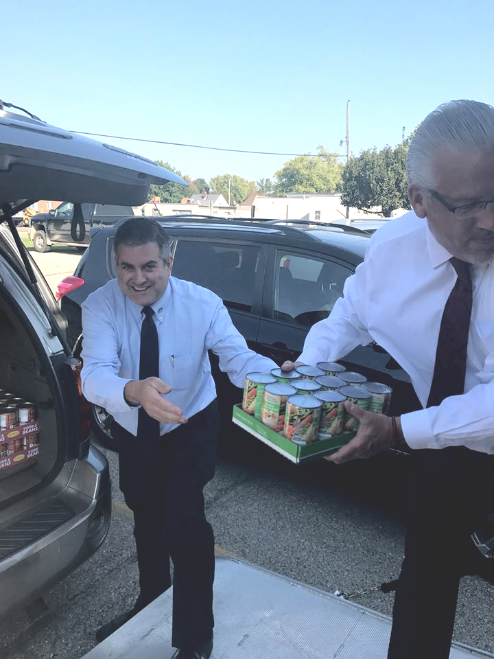 Two people in shirts and ties unload a case of canned food from the trunk of a car parked outdoors.