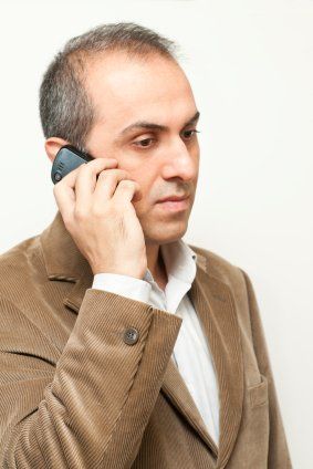 A man in a brown corduroy jacket and white shirt holds a phone to his ear against a plain white background.
