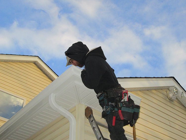 Person in black jacket working on a white gutter ladder beside a yellow house under a blue sky