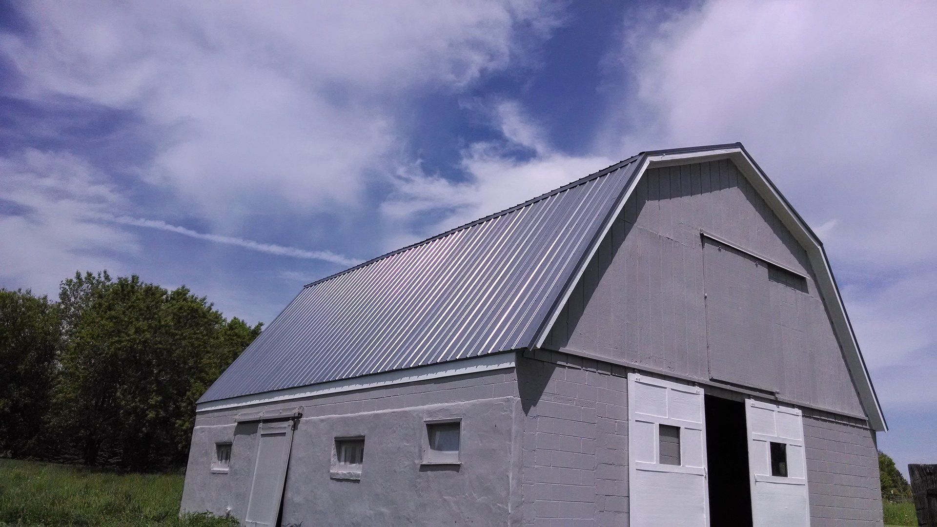 Gray barn with a steep metal roof under a cloudy sky, viewed from the side.