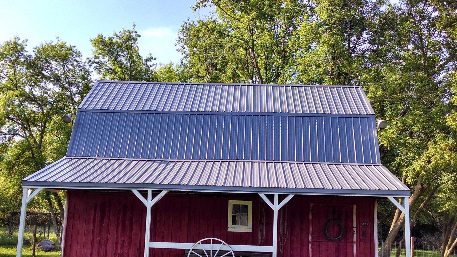 Red barn with a metal roof and front porch, surrounded by green trees under a clear sky