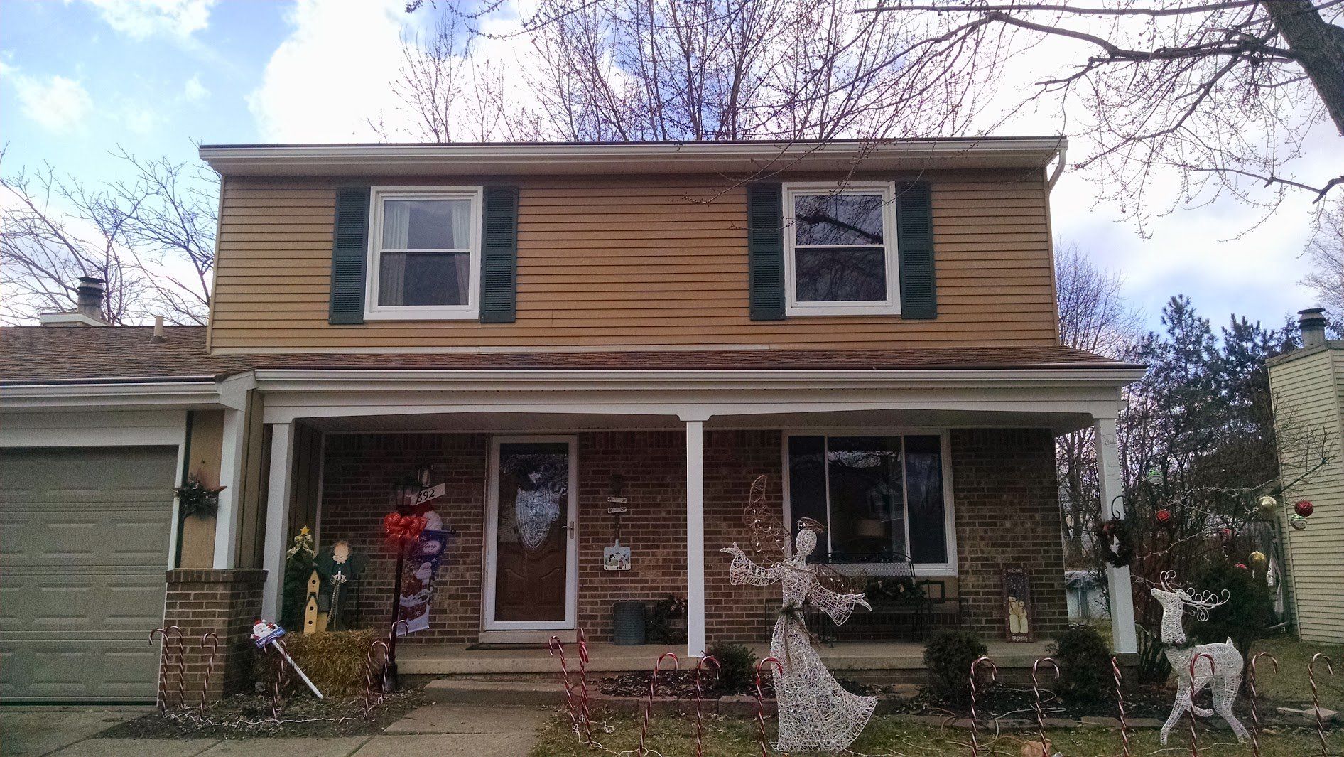 Two-story tan house with a front porch, flower decorations, and a small lawn under a cloudy sky